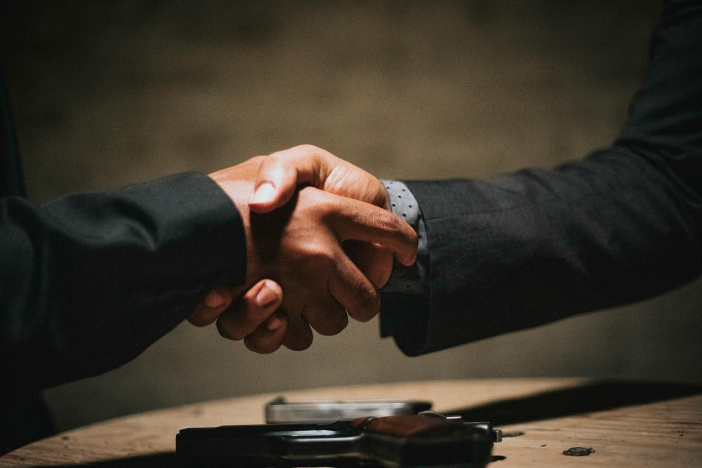 Two business executives smiling and shaking hands in front of a glass window overlooking the city, representing strategic partnerships and enterprise growth